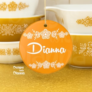 May include: A round, orange ceramic ornament with the name "Dianna" in white script. The ornament is decorated with white floral designs and the text "Designs with Dianna". The background features yellow and white kitchenware.