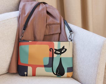 May include: A rectangular crossbody bag featuring a black cat illustration against a background of overlapping geometric shapes in orange, teal, and cream. The bag has a black zip closure and a thin black shoulder strap with silver metal hardware.