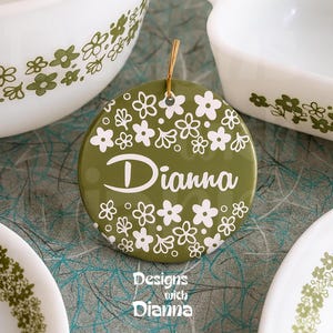 May include: A round, olive green ornament with the name "Dianna" in white script, surrounded by white floral designs. The ornament hangs by a gold string. The text "Designs with Dianna" is at the bottom. White dishware with green floral patterns is in the background.