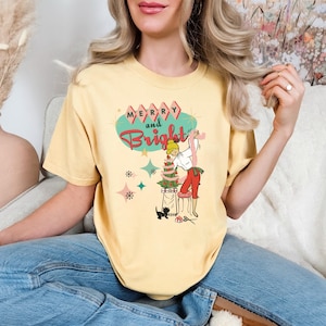 May include: Yellow t-shirt with a retro Christmas design. The graphic features the words "Merry and Bright" in red and turquoise, along with a cartoon illustration of a person decorating a Christmas tree. A black cat sits nearby.