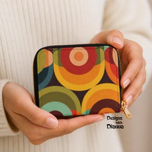 May include: A small square zippered wallet featuring a repeating pattern of concentric circles in shades of orange, yellow, olive green, and dark blue on a black background. The wallet has a gold-toned metal zipper pull and a black fabric border.