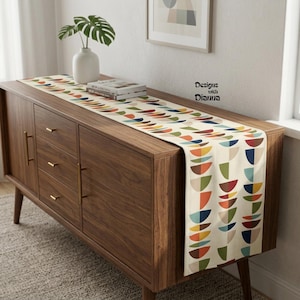 May include: A table runner featuring a repeating pattern of multicolored semi-circles in shades of blue, orange, yellow, and green on a cream background. The text Designs with Dianna is printed on the surface. The runner rests on a wooden cabinet.