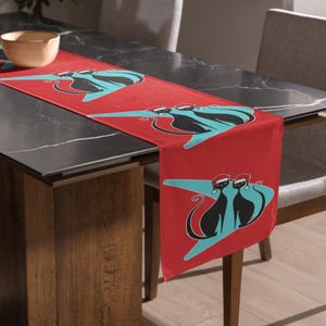 May include: Red table runner featuring a repeating pattern of black cat silhouettes wearing Santa hats on a turquoise starburst design. The runner is displayed on a dark gray table.