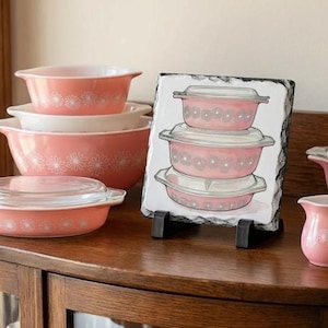 May include: A collection of pink and white floral patterned Pyrex bowls and dishes. The set includes various sizes of bowls with lids, and a decorative slate featuring a similar design. The bowls are arranged on a wooden surface.