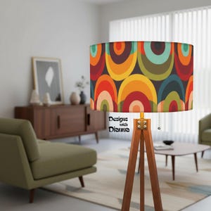 May include: A retro-style floor lamp with a wooden tripod base and a cylindrical lampshade. The lampshade features a colorful pattern of overlapping arcs in shades of red, orange, yellow, green, and blue. The text "Designs with Dianna" is printed on the lampshade.