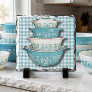 May include: A decorative slate tile featuring a stack of vintage turquoise and white mixing bowls with a retro design. The bowls are set against a blue and white checkered background, supported by a black stand.