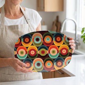 May include: Oval serving platter with a retro design. The platter features a repeating pattern of colorful concentric circles in shades of red, orange, yellow, green, and blue. The platter is held by a person wearing a white shirt and a beige apron.