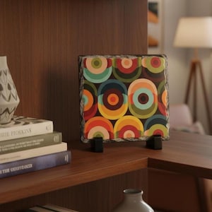 May include: A square decorative slate tile with a retro geometric design featuring overlapping circles in shades of orange, red, green, and blue. The tile is supported by two small black stands and sits on a wooden surface.