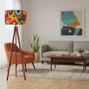 May include: A mid-century modern floor lamp with a colorful, retro-style lampshade. The shade features a pattern of overlapping arcs in shades of red, orange, yellow, green, and blue. The lamp has a dark wood tripod base and stands near an orange armchair.