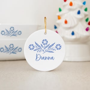May include: A white ceramic Christmas ornament with a blue floral design and the name "Dianna" in script. The ornament hangs from a gold string. In the background are matching casserole dishes and a white Christmas tree.