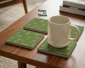 May include: A speckled cream-colored ceramic mug sits atop a square green coaster featuring a gold starburst pattern. Two additional matching square coasters are visible nearby. The text Designs with Dianna is printed above the coasters.