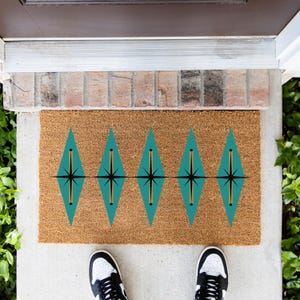 May include: A rectangular natural coir doormat featuring a repeating pattern of five teal diamond shapes. Each diamond contains a centered black starburst design with a vertical gold line, creating a geometric motif across the brown fiber surface.