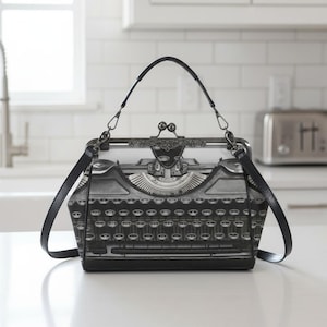 May include: A black and white handbag featuring a vintage typewriter design. The bag has a clasp closure, a top handle, and a detachable shoulder strap. The typewriter image is detailed, with keys and mechanical elements.