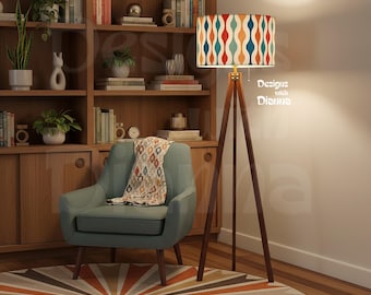 May include: A floor lamp with a tripod wooden base and a cylindrical shade featuring a repeating wavy pattern in orange, teal, and cream. The lamp stands next to a light blue armchair with a patterned throw blanket and a wooden bookshelf.