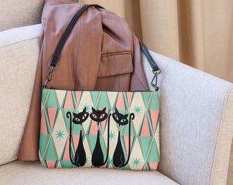May include: A rectangular crossbody bag featuring a repeating geometric pattern of teal and pink diamonds with small starburst accents. The front of the bag displays three stylized black cat silhouettes with long, curved tails and pointed ears.