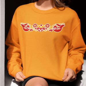 May include: A bright orange crewneck sweatshirt featuring a central graphic design of two stylized red birds facing outward, separated by red and yellow floral motifs, all outlined in white, with long sleeves and a ribbed collar and cuffs.