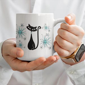 May include: A white ceramic mug featuring a stylized black cat illustration with a long, curved tail. The design includes several teal and black starburst patterns surrounding the cat, creating a graphic, mid-century inspired aesthetic.