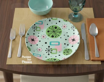 May include: A round dinner plate with a light mint green background featuring a pattern of black starbursts, pink circles, and green geometric shapes. The plate is set with two silver forks, a silver knife, a silver spoon, and a glass.