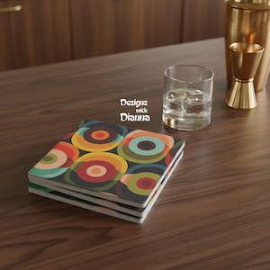 May include: A stack of square coasters with a retro design of colorful concentric circles in shades of orange, red, green, and blue on a dark background. The coasters are on a wooden surface, with a glass of water and bar tools in the background. Text on the coasters reads "Designs with Dianna."