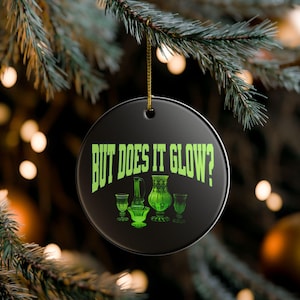 May include: A round black ceramic ornament featuring the lime green text "BUT DOES IT GLOW?" above a graphic illustration of four pieces of green glassware, including a pitcher, a bowl, and two stemmed glasses, suspended by a gold string.