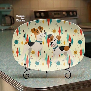 May include: An oval ceramic platter featuring a repeating pattern of two Jack Russell terriers, red and teal diamonds, and starburst shapes on a cream background. The platter is displayed on a black metal stand. Text reads Designs with Dianna.