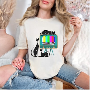 May include: Cream-colored t-shirt featuring a retro television design with a colorful test pattern and three black cat silhouettes. The cats are in various poses, adding a playful touch to the design. The shirt is made of a soft material.