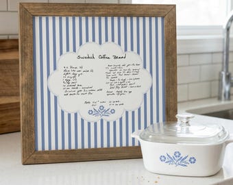 May include: A framed print featuring a blue and white striped background with a central cloud-shaped label containing a handwritten recipe for Swedish Coffee Bread and a small blue floral illustration, next to a white ceramic dish with a glass lid.