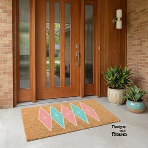 May include: A rectangular coir doormat with a retro design. The mat features a repeating pattern of pink and turquoise diamond shapes. The text "Designs with Dianna" is in the lower right corner. The mat is placed in front of a wooden door.