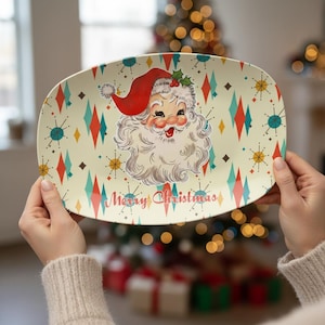May include: An oval-shaped serving platter featuring a central illustration of a smiling Santa Claus wearing a red hat. The background is cream-colored with a repeating pattern of teal and red diamonds and starbursts, with the text Merry Christmas.