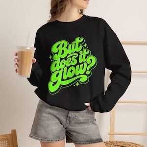 May include: A black long-sleeve sweatshirt featuring the bright neon green text "But does it glow?" in a stylized, rounded font. The graphic includes small star accents surrounding the lettering, which is centered on the front of the garment.