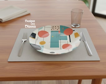 May include: A round ceramic plate featuring a geometric pattern of teal rectangles, coral circles, yellow dots, and black lines. The text 'Designs with Dianna' is printed in white. A silver fork and knife are placed on a gray placemat with a glass.
