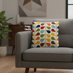 May include: Square throw pillow with a retro teardrop pattern in shades of red, blue, yellow, orange, green, and brown on a cream background. The pillow is on a gray sofa. The text "Designs with Dianna" is visible.