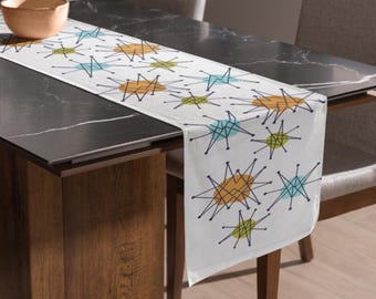 May include: A white table runner featuring a repeating pattern of black starburst lines overlaid on solid circular shapes in shades of teal, mustard yellow, and lime green. The fabric has a rectangular shape with finished edges and a smooth texture.