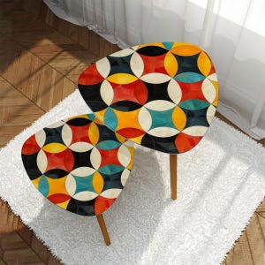 May include: Two retro-style side tables with a colorful geometric pattern. The tabletops feature a design of overlapping circles in red, yellow, black, and teal. The tables have wooden legs and are set on a white rug.