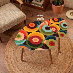 May include: Two nesting tables with a colorful pattern of overlapping circles in shades of orange, yellow, red, green, and teal. The tables feature three light-colored wooden legs each and are marked with the text Designs with Dianna.