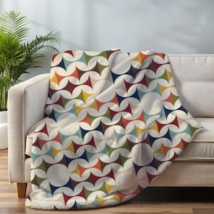 May include: A soft, cream-colored throw blanket with a repeating pattern of colorful starburst shapes in shades of red, orange, yellow, green, blue, and brown. The blanket is draped over a beige sofa, creating a cozy and inviting aesthetic.