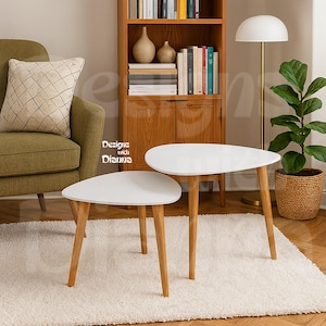 May include: Two white, teardrop-shaped side tables with light wood legs. The tables are on a white rug, with a green armchair and wooden cabinet in the background. The text "Designs with Dianna" is visible.