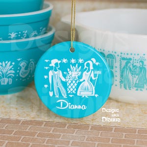 May include: A round, turquoise ceramic ornament with a gold string for hanging. The ornament features a white illustration of a couple holding a basket of flowers. The name "Dianna" is written in white script. The words "Designs with Dianna" are also visible.