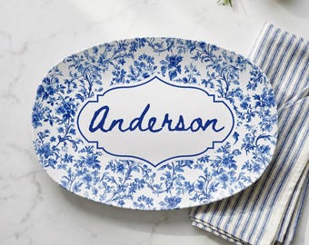 May include: An oval serving platter featuring a blue and white floral pattern. The center of the platter has a white decorative frame containing the name Anderson written in blue script. The platter is positioned next to a blue and white striped cloth.