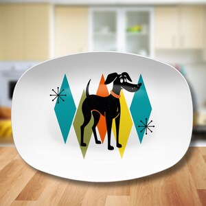 May include: White oval serving platter with a black dog silhouette standing on a background of colorful geometric shapes. The dog is looking to the right and has a collar. The geometric shapes are in shades of blue, green, yellow, and orange.