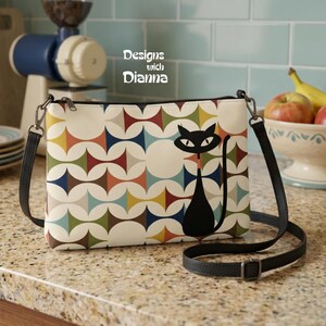 May include: A rectangular crossbody bag featuring a repeating geometric pattern of circles and diamonds in red, blue, yellow, and green on a cream background. A black cat silhouette is printed on the right side. The text Designs with Dianna is visible.