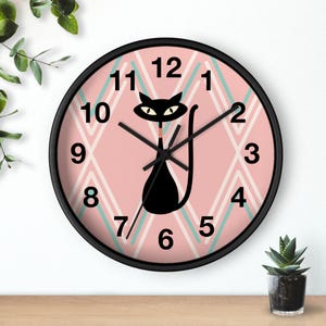May include: Black and pink wall clock with a cat design. The clock face features a pink background with a diamond pattern in white and teal. The black cat silhouette has large eyes and a long tail. The clock is in a black frame.