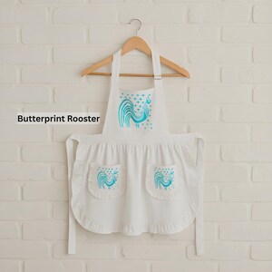 May include: A white apron featuring a teal rooster graphic on the chest and two front pockets, each decorated with a matching teal rooster print. The apron includes a ruffled hem and is displayed on a wooden hanger against a white brick wall.