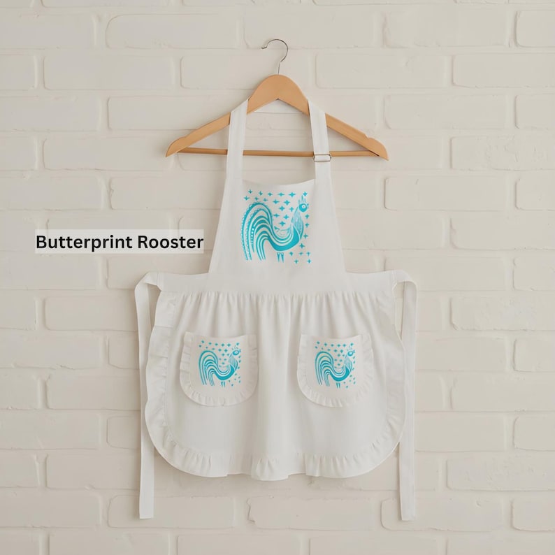 May include: A white apron featuring a teal rooster graphic on the chest and two front pockets, each decorated with a matching teal rooster print. The apron includes a ruffled hem and is displayed on a wooden hanger against a white brick wall.