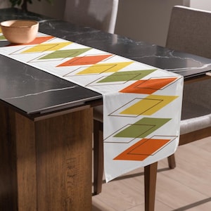 May include: A white table runner featuring a repeating geometric pattern of orange, yellow, and olive green diamonds outlined in thin black lines. The runner is draped across a dark surface, with a small, plain, light-colored bowl placed on one end.