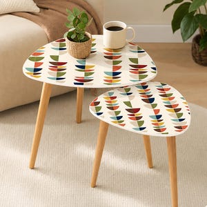 May include: Two nesting tables with light wood legs and white triangular tops featuring a repeating pattern of multicolored semicircles in red, green, blue, and yellow. A small potted green plant and a white ribbed mug sit on the larger table.