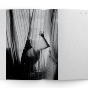May include: A black and white photograph of a person standing behind a sheer curtain. The person's hand is raised, and their body is partially obscured by the curtain.
