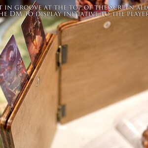 May include: A wooden game master screen with a built-in groove at the top for displaying initiative cards. The screen is open, revealing a brown interior. Two cards are visible in the groove, one with a dragon and the other with a fantasy scene.