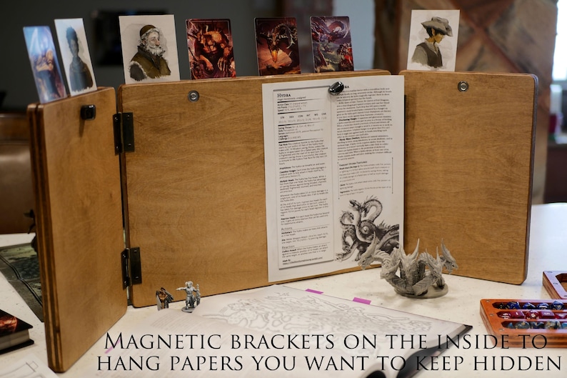May include: A wooden tabletop gaming screen with magnetic brackets on the inside to hold papers. The screen is folded open to reveal a printed sheet of paper with the word "Heroes" at the top. There are also several small images attached to the screen with magnets.