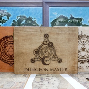 May include: Three wooden boxes with engraved designs. The center box has a large, intricate design with a five-pointed star, a crescent moon, and a 20-sided die. The text "DUNGEON MASTER" is engraved below the design. The box on the left has a circular design with a 20-sided die. The box on the right has a dragon design with a 20-sided die. The text "ON MASTER" is engraved below the design.